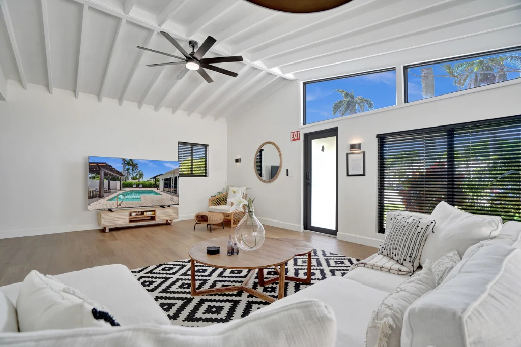 Bright living room with white vaulted ceiling, large windows, sectional sofa, and TV displaying a pool view