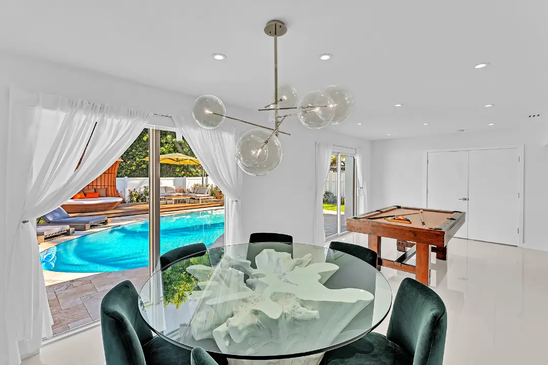 Dining room with glass table, modern chandelier, and pool table overlooking backyard pool.