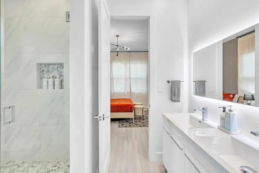 Bright bathroom with dual-sink vanity and walk-in shower leading to bedroom with orange bedding.