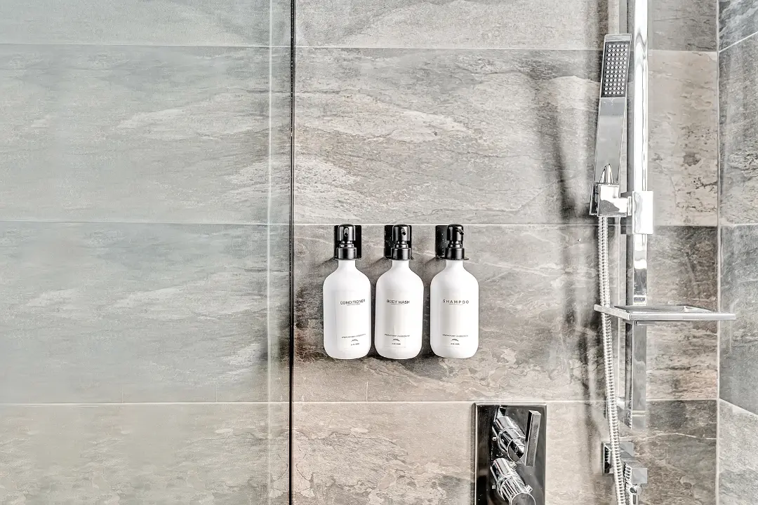 Wall-mounted shampoo, conditioner, and body wash beside chrome shower fixtures in a rental bathroom.